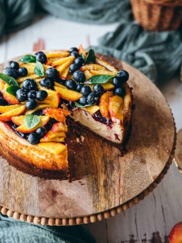 close up image at side angle showing blueberry peach cheesecake on a rustic wooden cake stand covered in fresh blueberries and sliced peach wedge with mint leaves for garnish