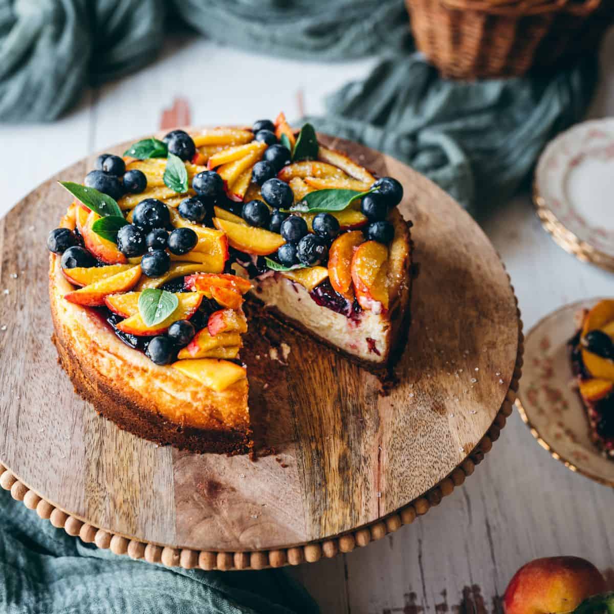 close up image at side angle showing blueberry peach cheesecake on a rustic wooden cake stand covered in fresh blueberries and sliced peach wedge with mint leaves for garnish