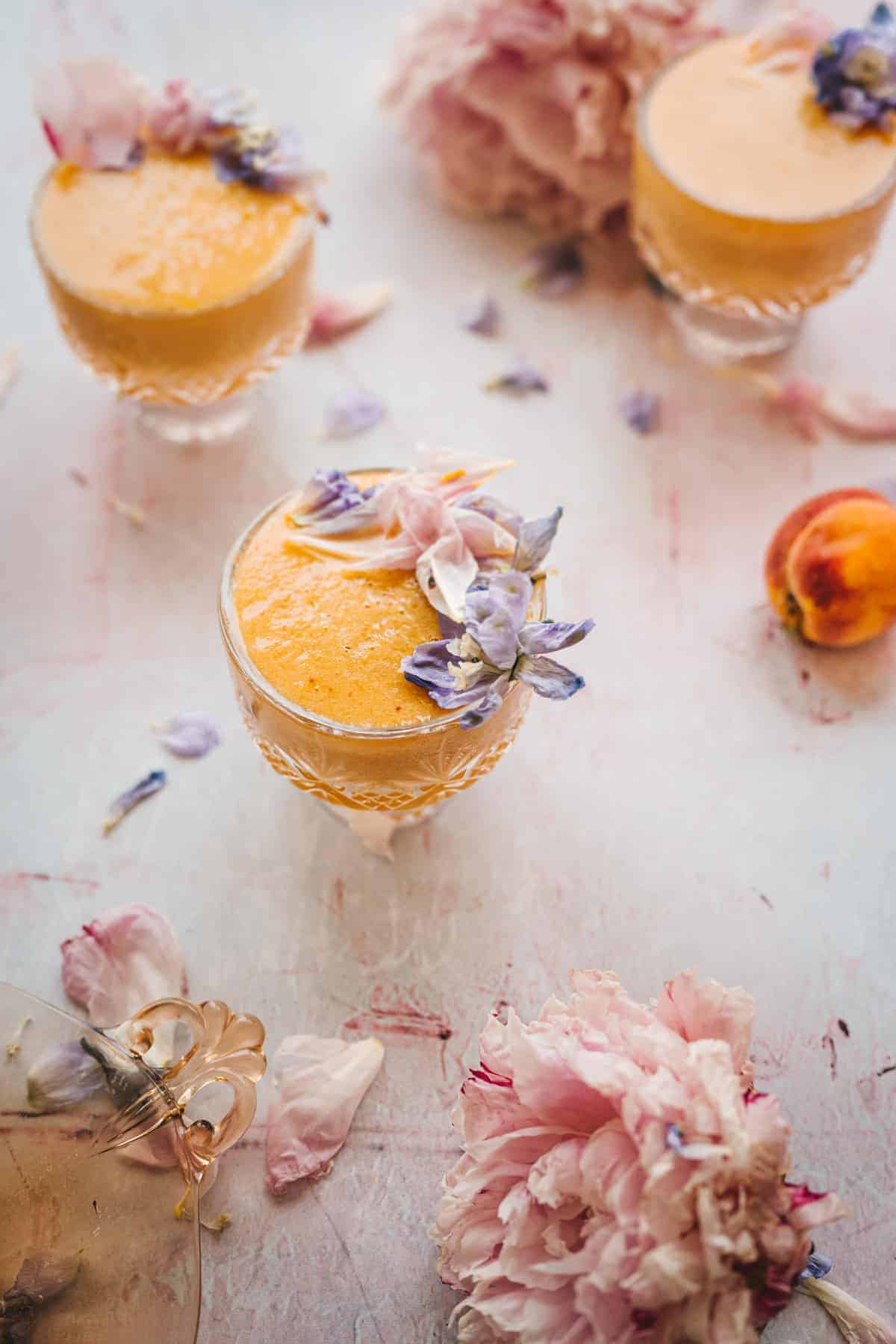 close up aged shot of a light pink marble surface with fallen petals all over and a frozen peach cocktail in crystal glass