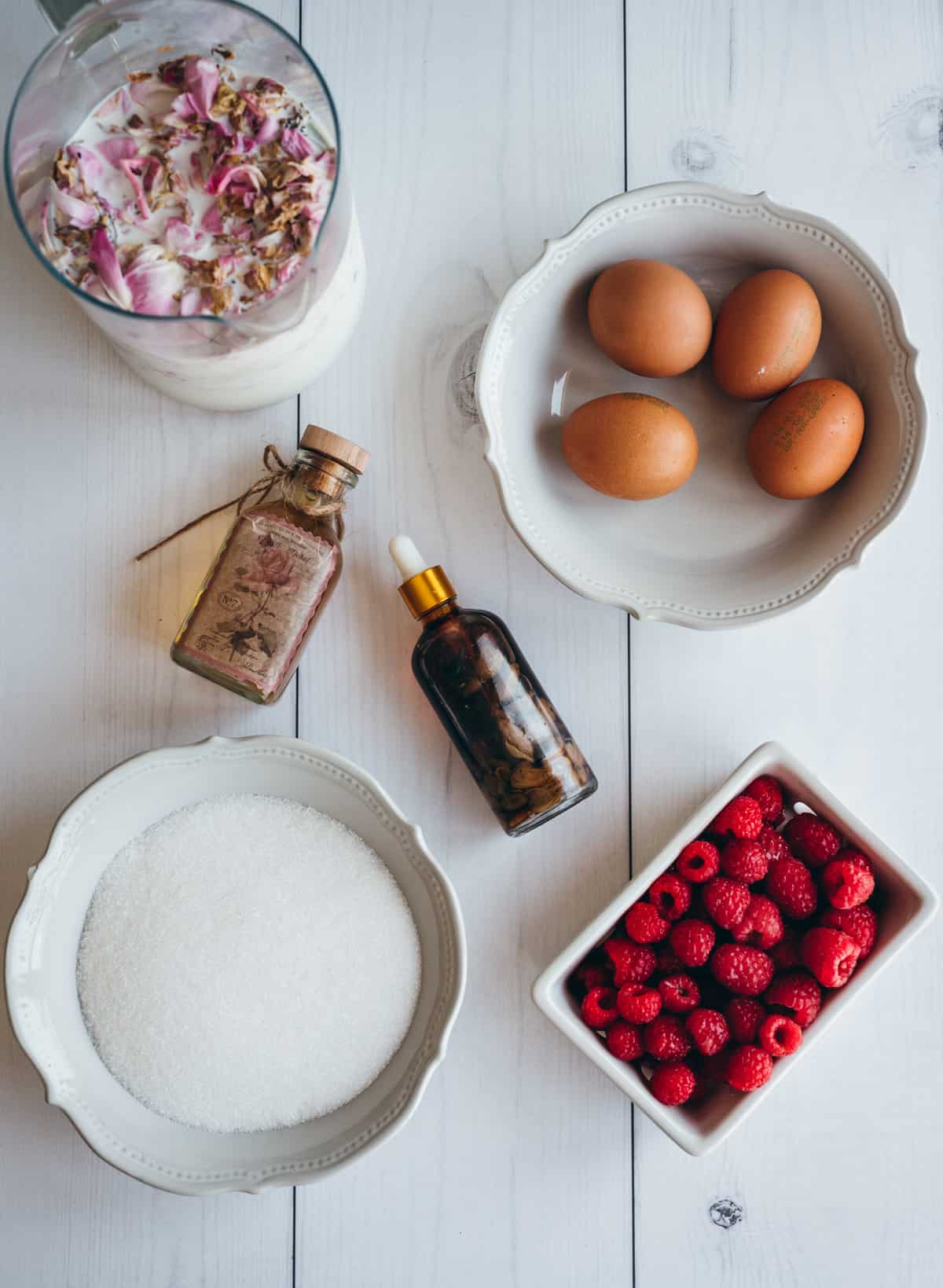 ingredients for rose ice cream including small doppler bottle of vanilla extract, a white bowl of sugar, a ceramic berry bowl filled with red raspberries, a white bowl with brown eggs and rose infused milk on a white rustic surface