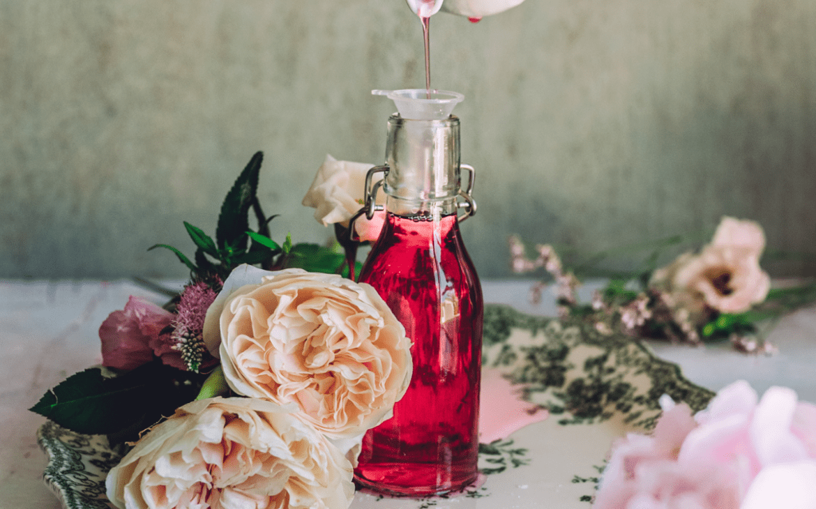 image of small bottle with pink rose syrup being poured into it in small funnel surrounded by roses on a vintage like background