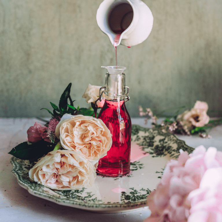 image of small bottle with pink rose syrup being poured into it in small funnel surrounded by roses on a vintage like background