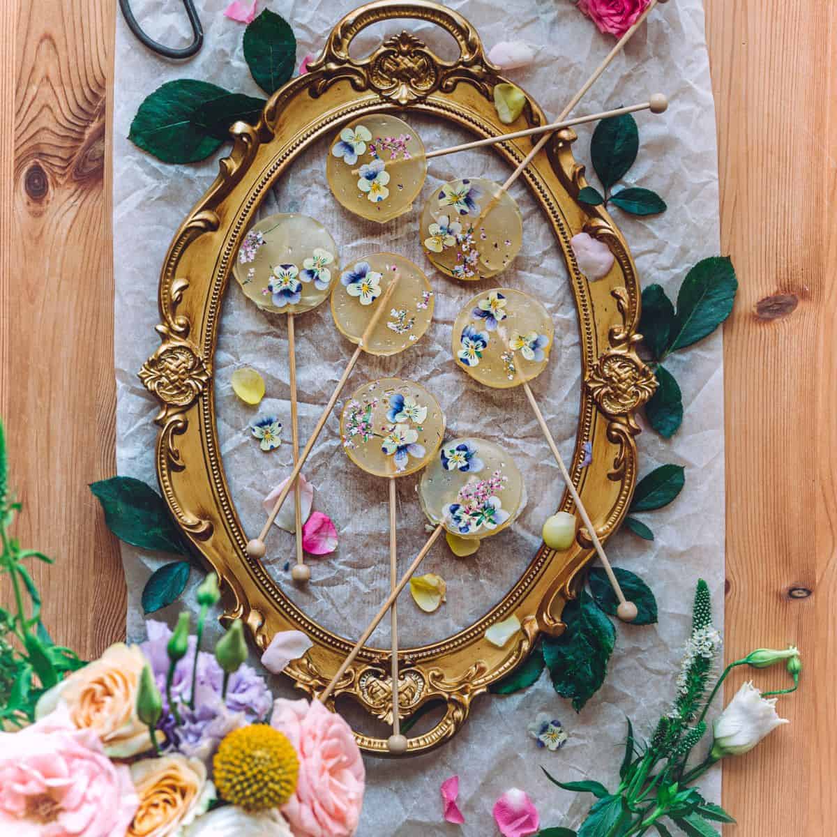 overhead image of golden frame with numerous edible flower lollipops inside of them on a rustic table surrounded by petals