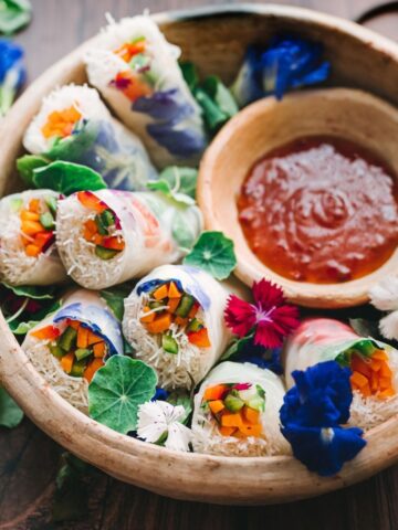 Edible flower spring rolls in a bowl at an angle so you can see vibrant vegetables and a small bowl of chili dipping sauce