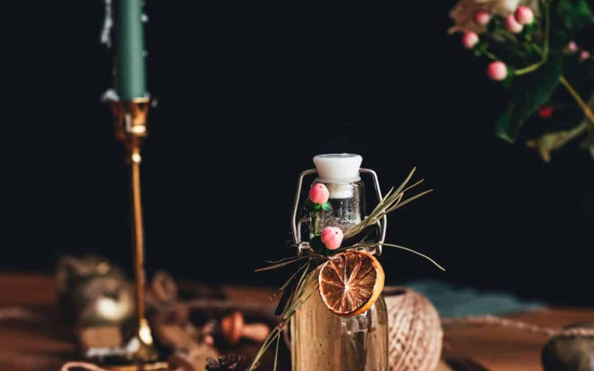 small glass bottle with pine needle syrup with twine tied around top holding a dried orange, pine needles and pink berries with a black background and sage green candle flickering in the background