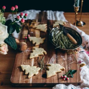 large rectangle cutting board with 6 light colored shortbread cookies shaped like pine christmas trees with an oblong copper bowl with pine needles and some pink berries scattered