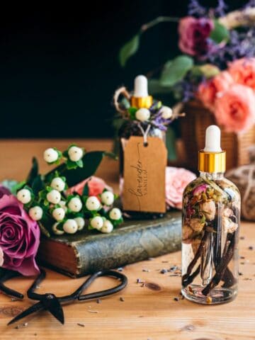 clear doppler bottles with gold embellishment filled with vanilla bean pods and rose buds and lavender flowers on top of a vintage book on a wood table surrounded by roses