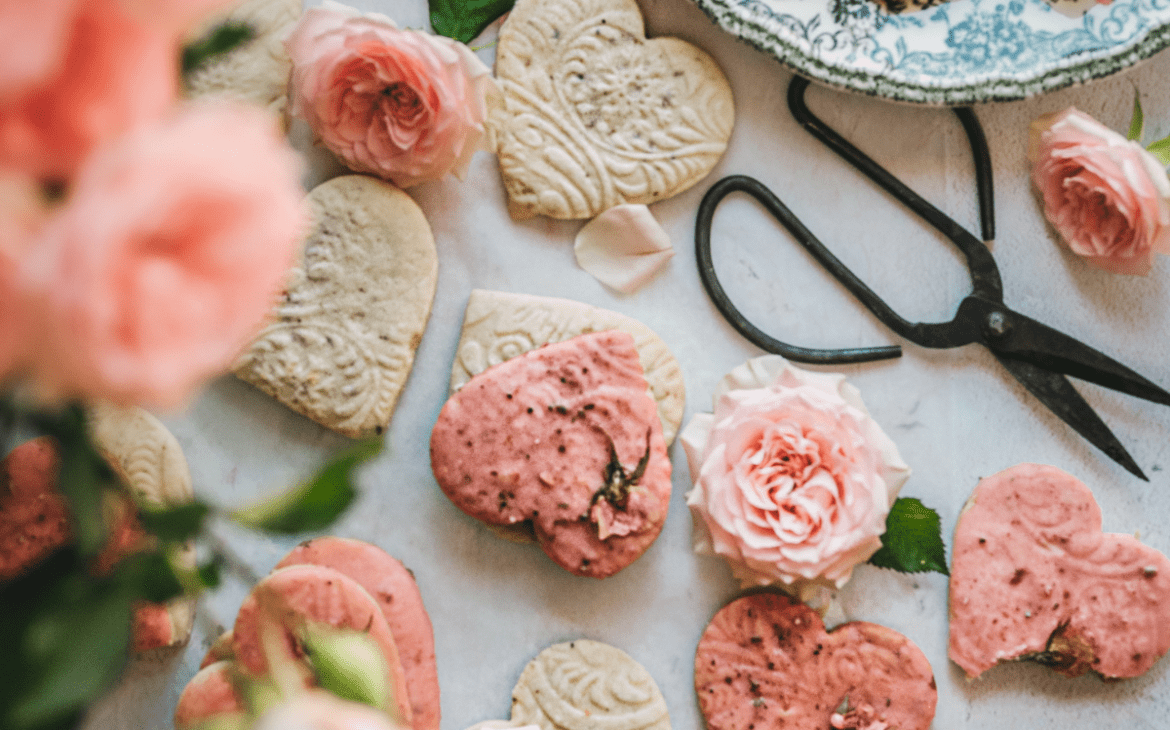 overhead shot of heart shaped cookies with embossed shapes and pink glaze next to a bouquet of pink roses and garden scissors