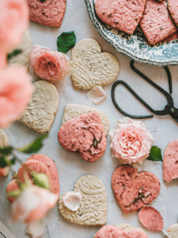overhead shot of heart shaped cookies with embossed shapes and pink glaze next to a bouquet of pink roses and garden scissors