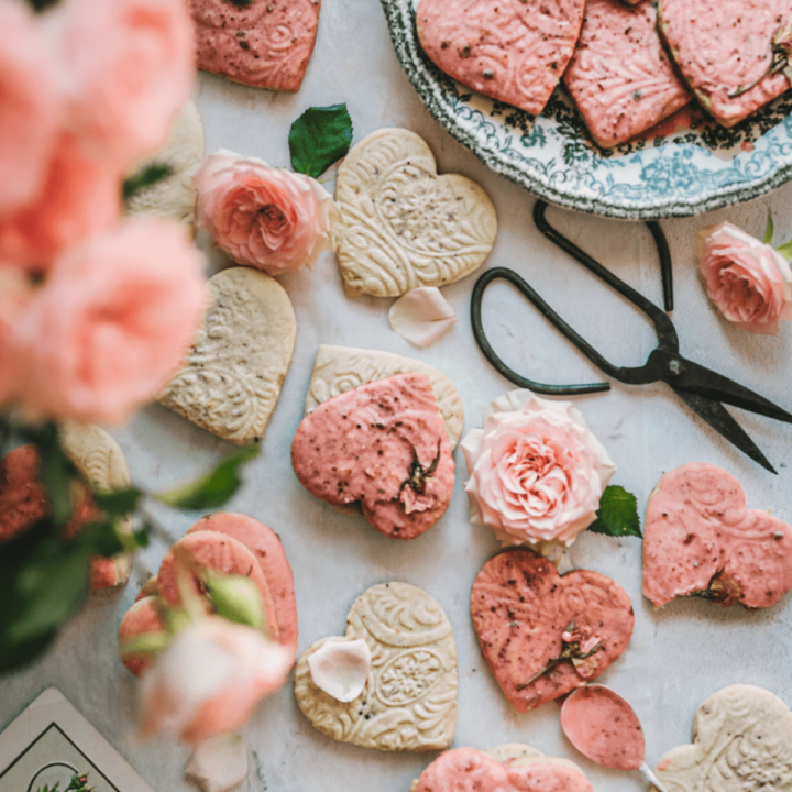 overhead shot of heart shaped cookies with embossed shapes and pink glaze next to a bouquet of pink roses and garden scissors