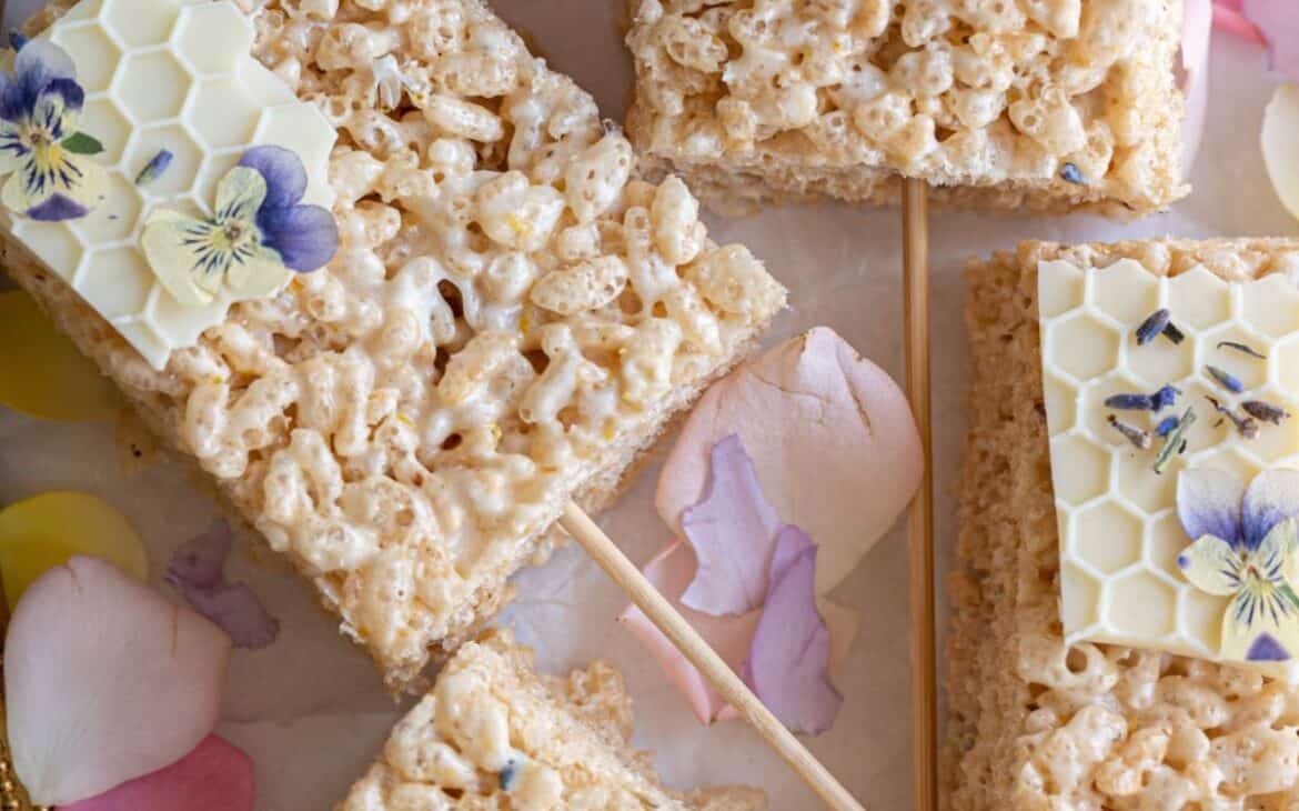 close up shot of perfectly rectangular rice krispy treat surrounded by pale pastel flower petals garnished with white chocolate honey comb and mini purple and yellow pansy flowers with wooden sticks in the center