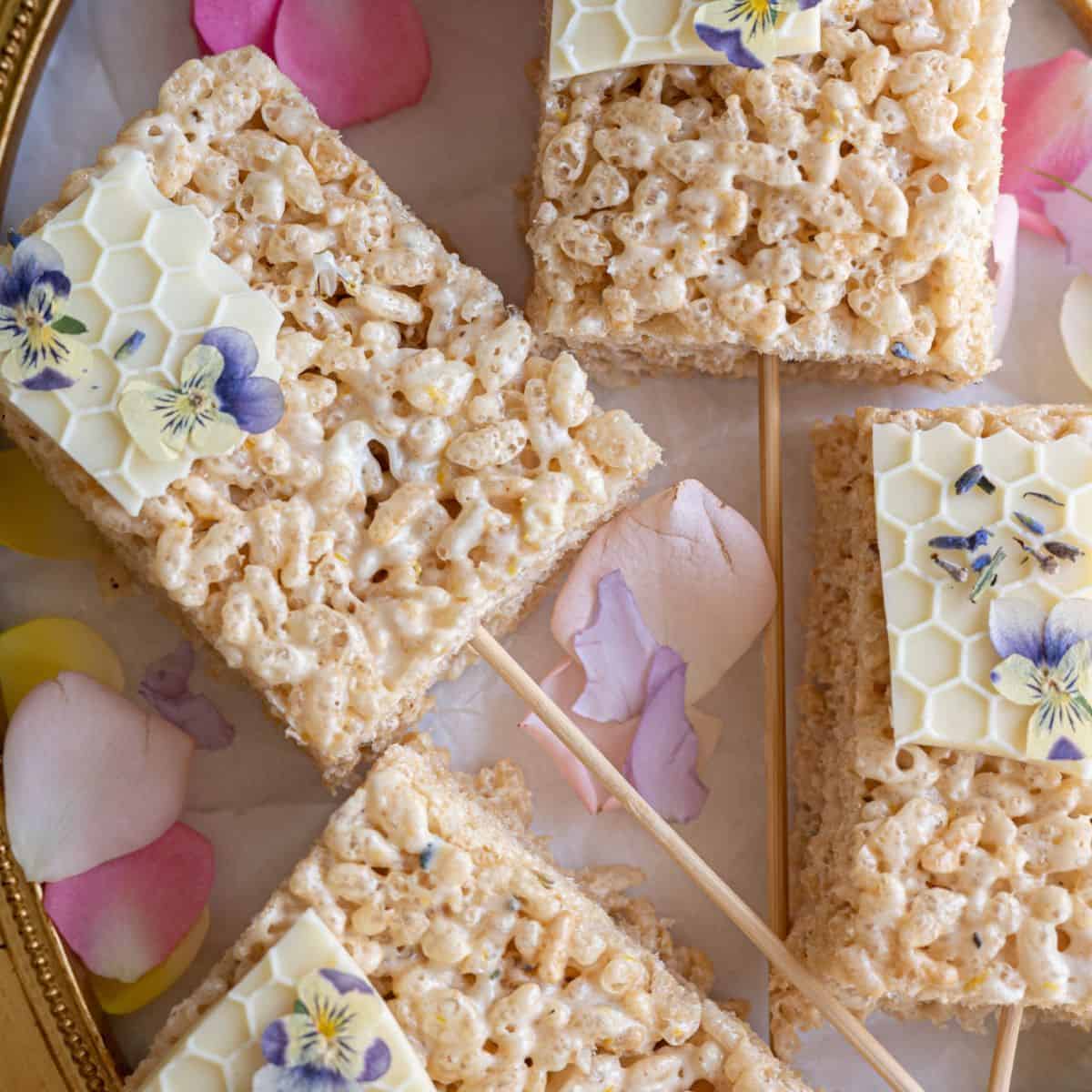 close up shot of perfectly rectangular rice krispy treat surrounded by pale pastel flower petals garnished with white chocolate honey comb and mini purple and yellow pansy flowers with wooden sticks in the center