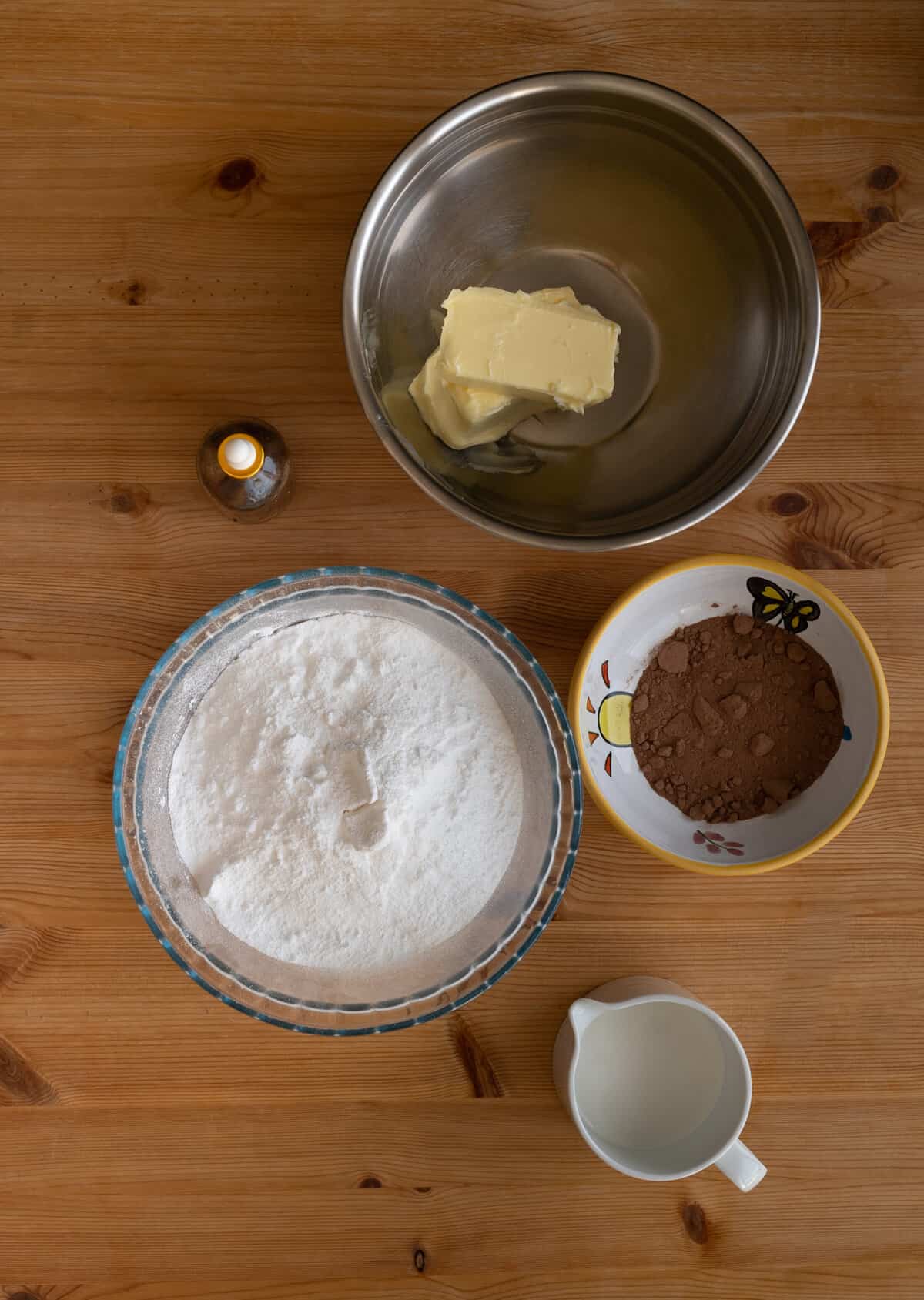 Ingredients in chocolate buttercream, stainless steel bowl with yellow butter, small bowl filled with confectionary sugar, and cocoa powder
