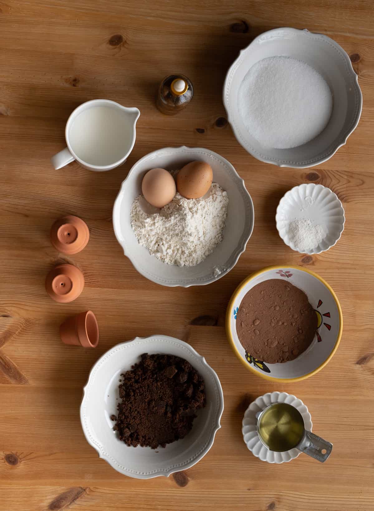 overhead shot of bowls of ingredients for dirt cupcakes, small porcelain bowl filled with milk, white bowl filled with flour and two eggs, small yellow bowl filled with cocoa powder, small bowl filled with baking soda and powder, and white porcelain bowl filled with white and brown sugar on a rustic brown tabletop