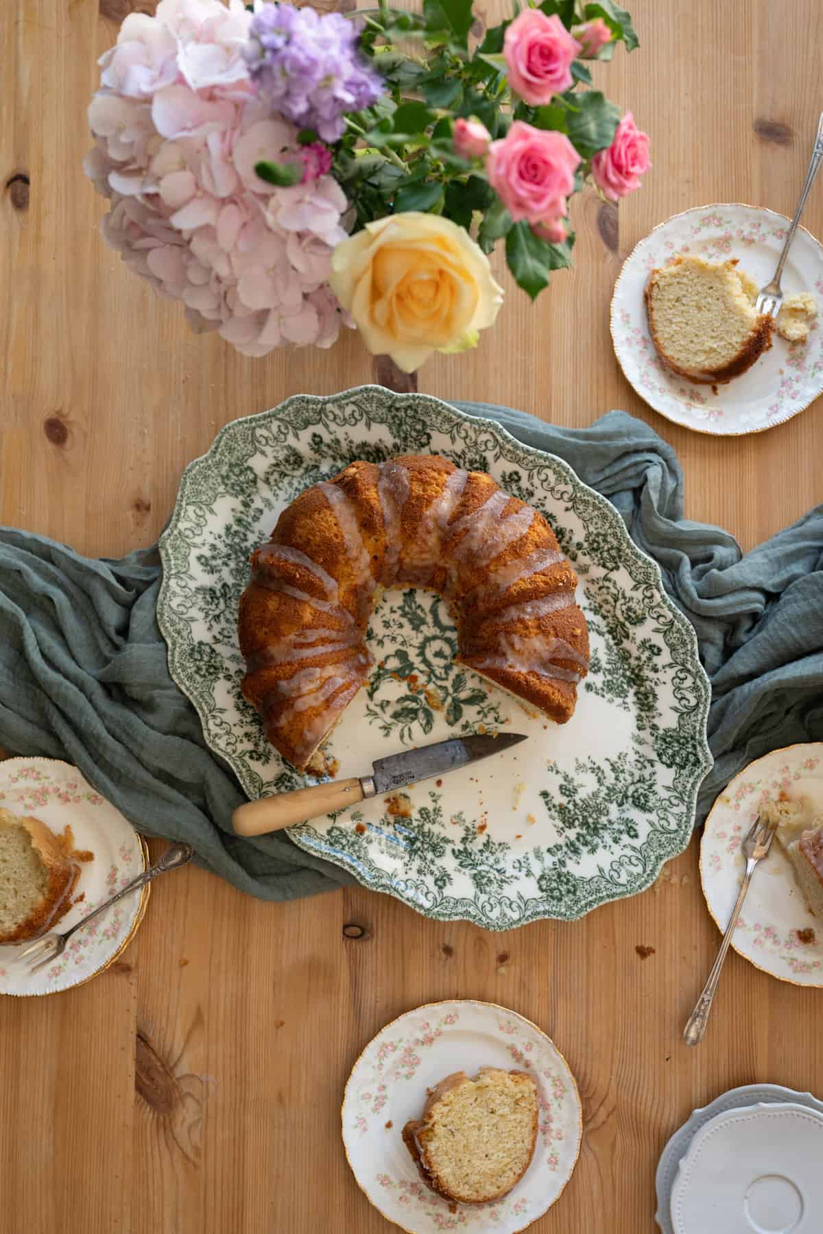 overhead image of green floral victorian serving dish with an ornate bundt cake drizzled with icing on top, with several floral rose dishes with slices of cake on top, and a bouquet of flowers on the left hand corner