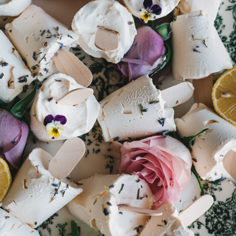 close up shot of light colored lavender lemon ice cream in cylinder shape with popsicle stick in many different directions, sprinkled with lavender buds, surrounded by lemon wedge, pink rose petals and pansies