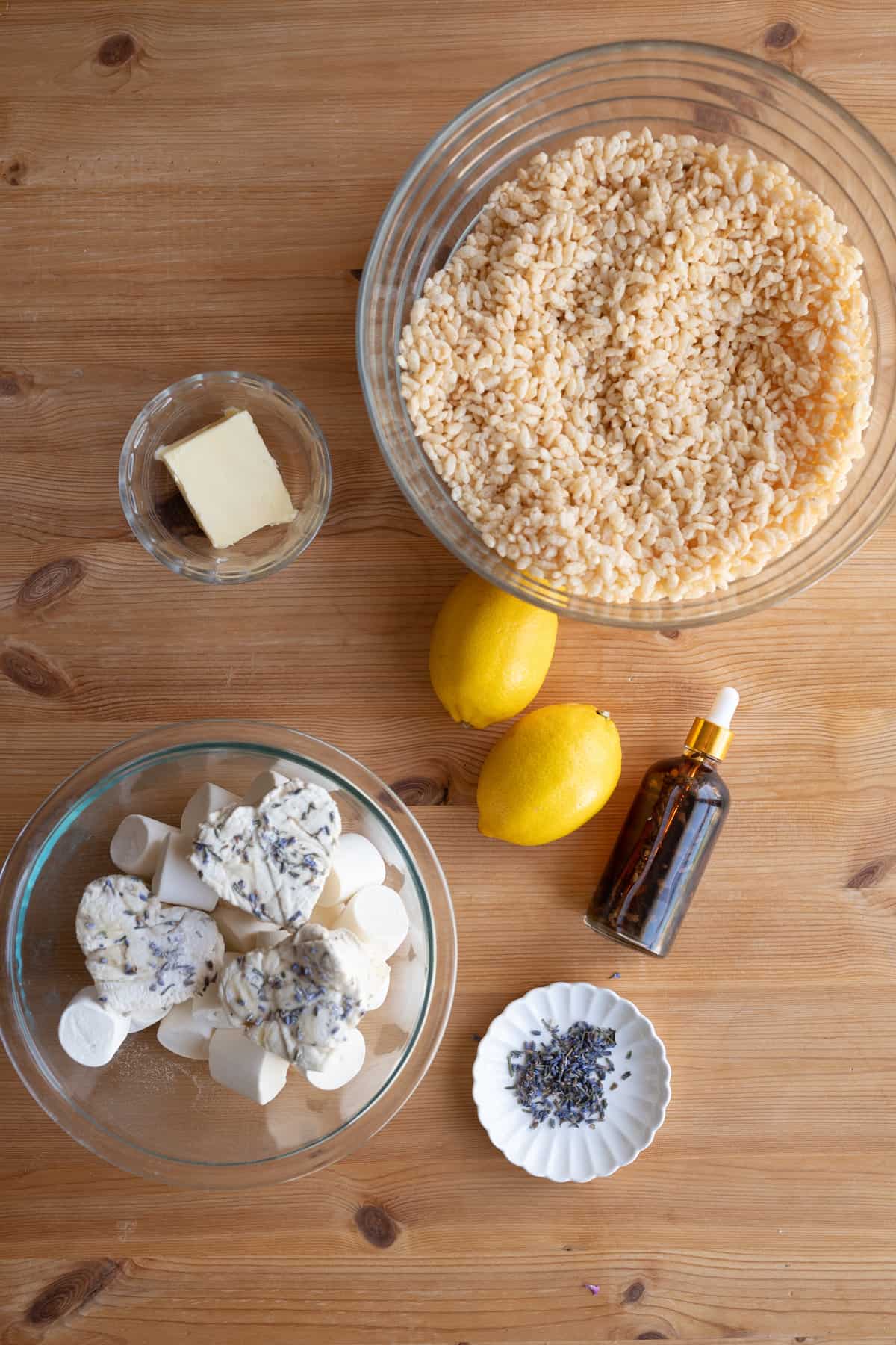 overhead image of ingredients for lemon lavender rice krispy treats, a bowl of rice krispy treats, a bowl of marshmallows, lemons, butter, lavender, and vanilla extract on a wooden rustic background