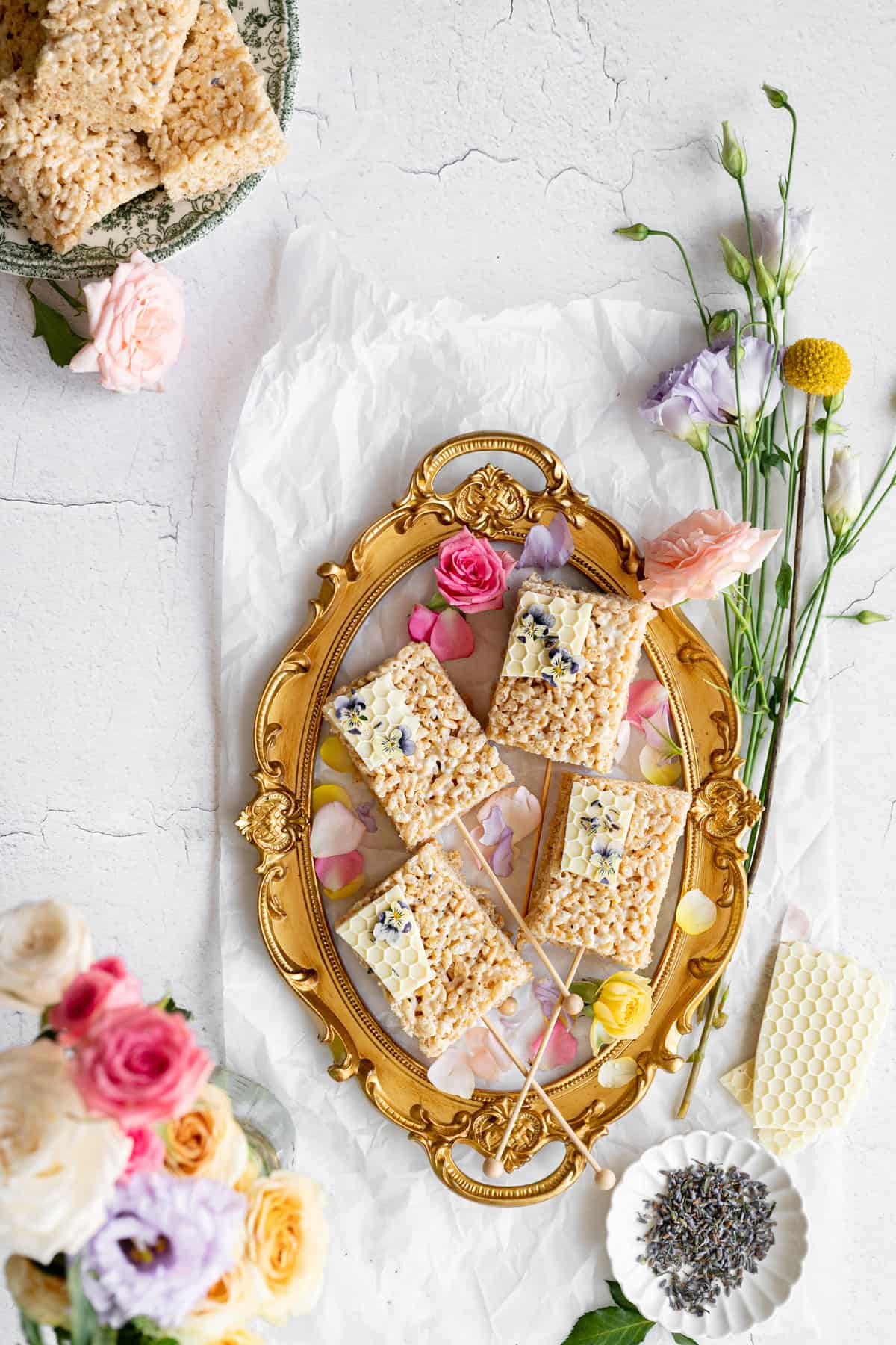 overhead image of vintage gold frame filled with rice krispy treats topped with white chocolate honey comb and pressed pansy flowers, next to a bouquet of pastel flowers on a rustic white concrete surface