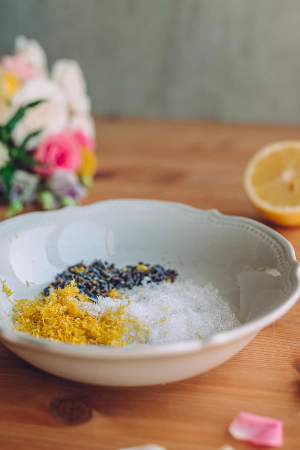 close up ornate white porcelain bowl filled with a scoop of white sugar, next to a scoop of lemon zest, next to a scoop of lavender buds