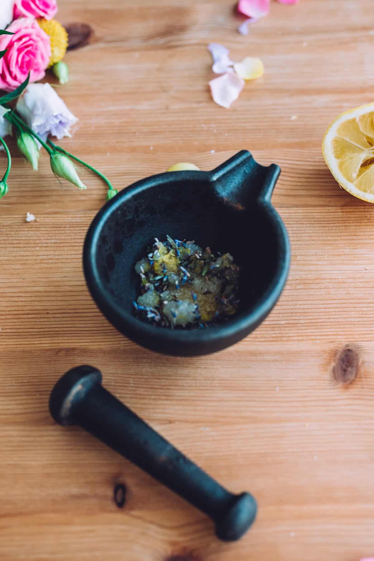 overhead shot of a cast iron mortar and pestle on a rustic table filled with lemon zest, lavender, and sugar next to a squeezed lemon