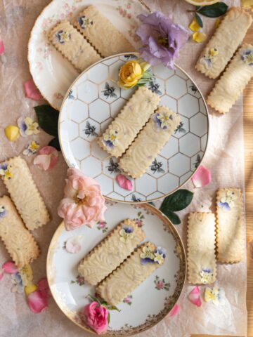 overhead image of 3 vintage tea saucer plates with rectangular shortbread cookies placed diagonally with dainty purple and yellow pansy flowers garnished on top