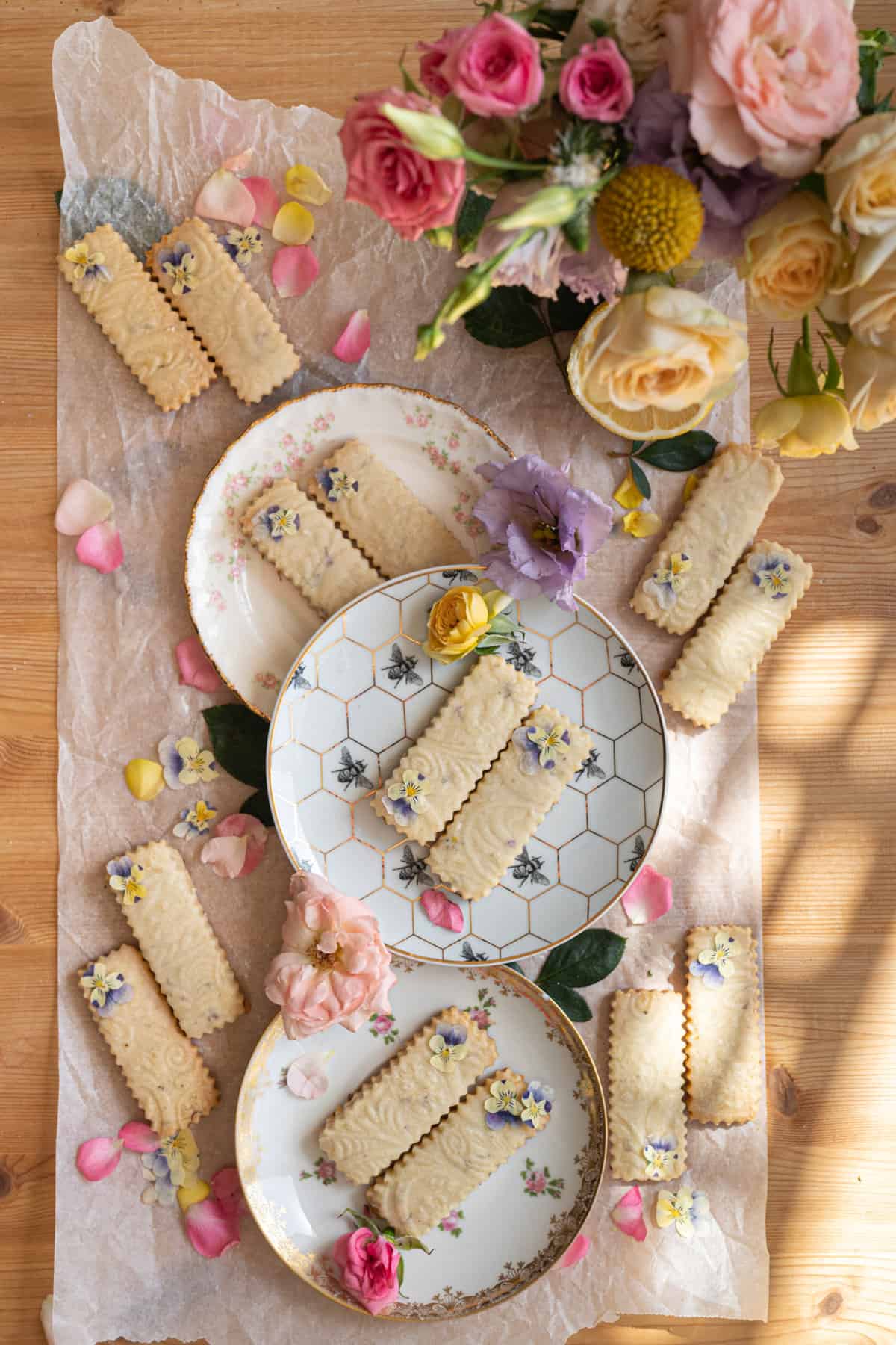 overhead image of 3 vintage tea saucer plates with rectangular shortbread cookies placed diagonally with dainty purple and yellow pansy flowers garnished on top with delicate dappled light coming in from the the corner