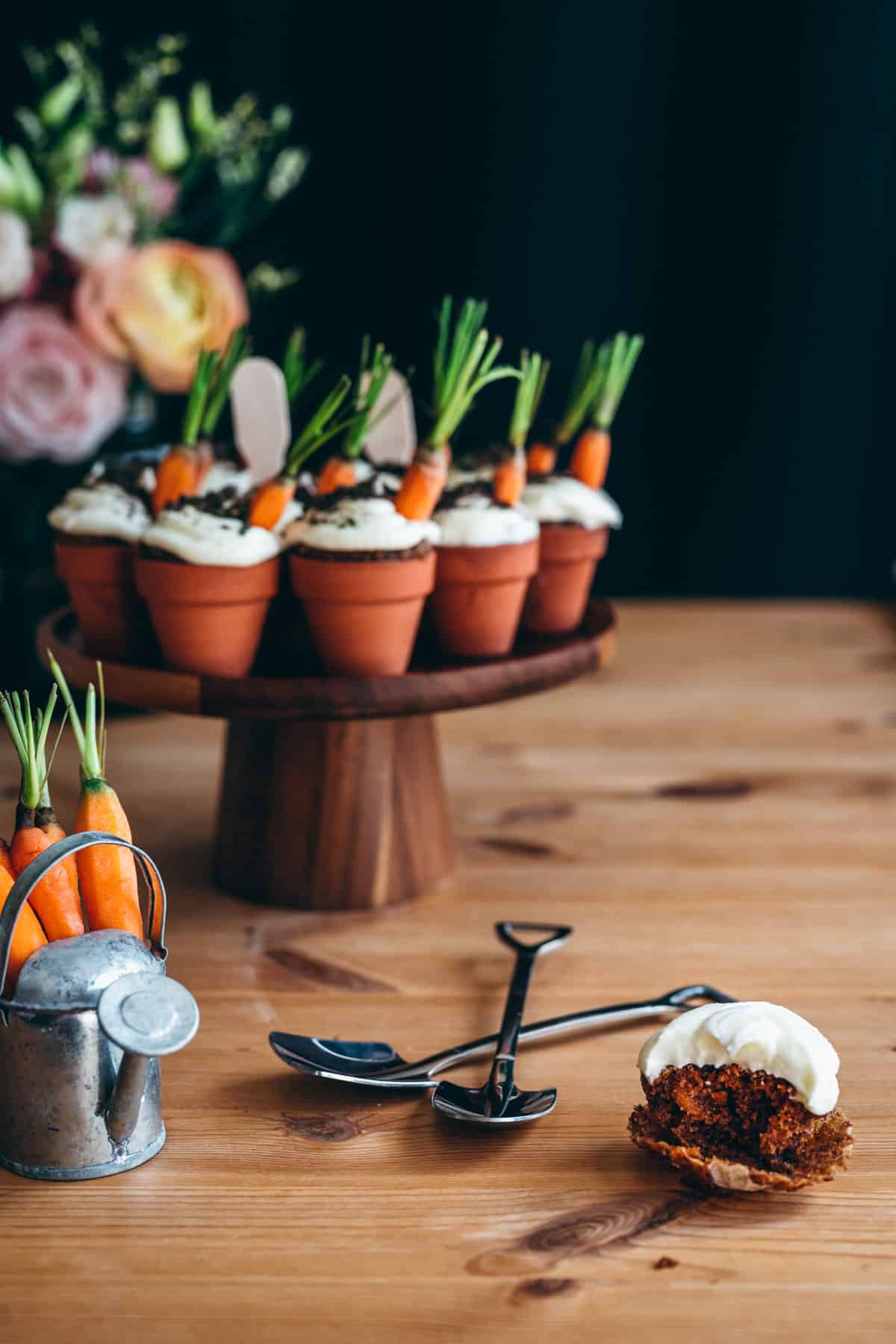 wooden cake stand topped with mini terracotta pots filled with carrot cake cupcakes, topped with cream cheese frosting, oreo "dirt" and mini carrots, next to a tiny watering can filled with baby carrots, and in front of orange ranunculus flowers