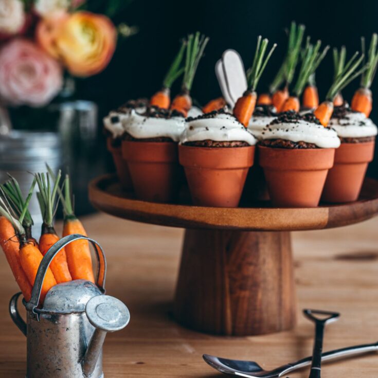 Mini carrot cake cupcakes in mini terracotta pots topped with mini carrots and oreo dirt on top a cake stand next to mini spoons and shovels