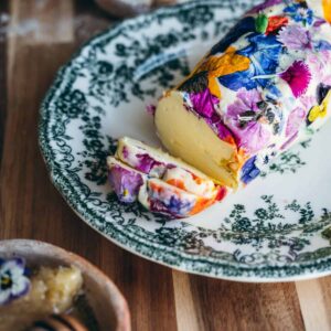 Close up macro image of compound butter log garnished with colorful edible flowers on a green Victorian Plate next to a small rustic dish filled with honey and in front of a rustic loaf of bread