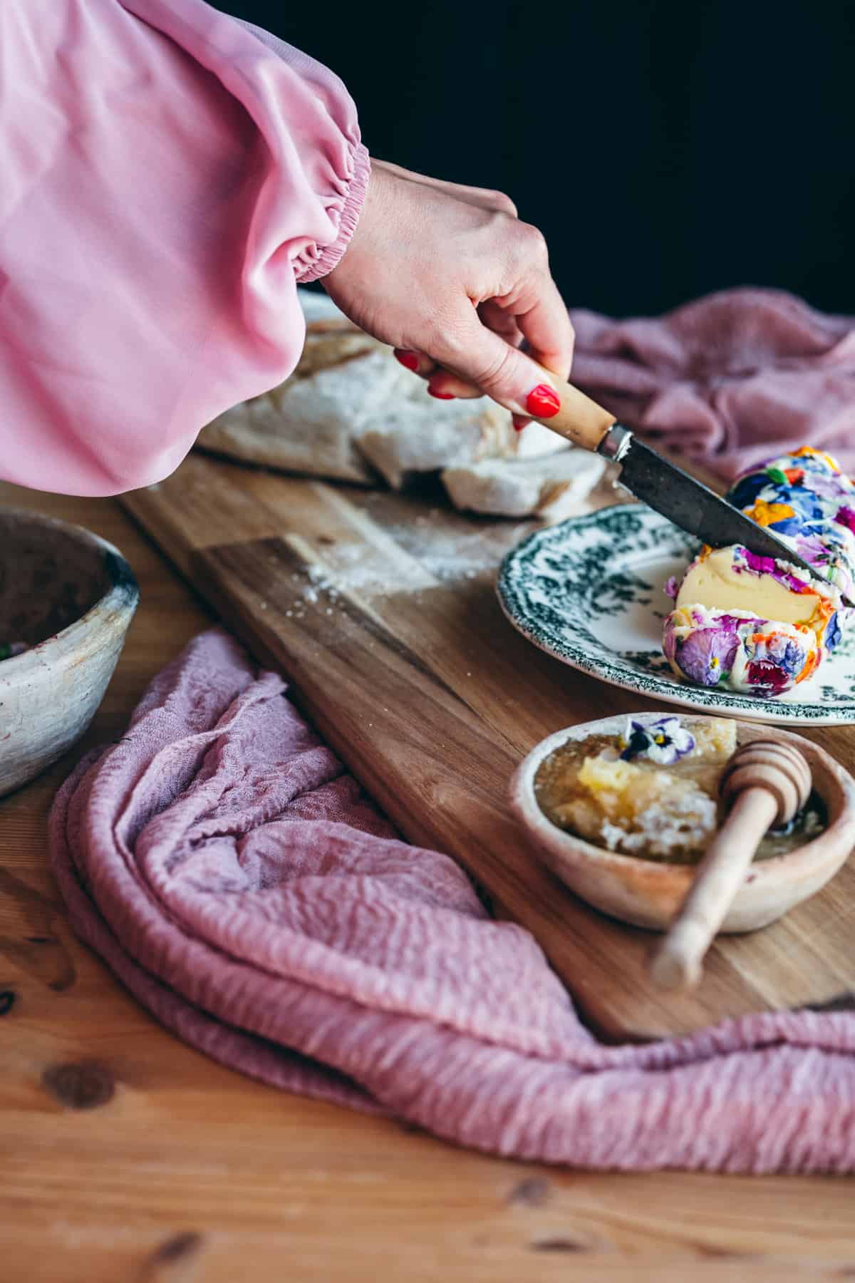 Woman's hand showing pink flowing sleeves and bright red finger nails holding a vintage knife cutting into flower compound butter, next to a loaf of bread and a small dish of honey with honey comb stick