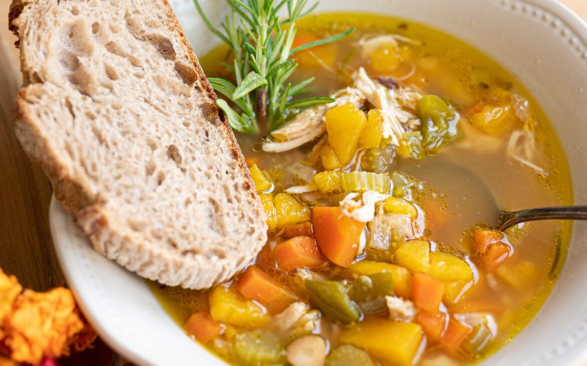 close up image looking into bowl of soup with chopped vegetable and slice of sourdough bread and herbs