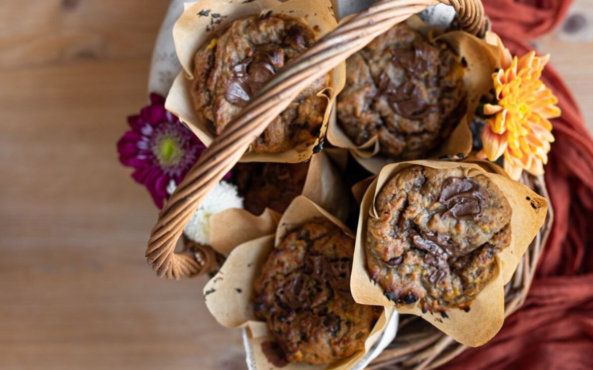 looking into basket of chocolalte chip covered pumpkin banana muffins on top of an orange cheesecloth next to a bouquet of autumn flowers