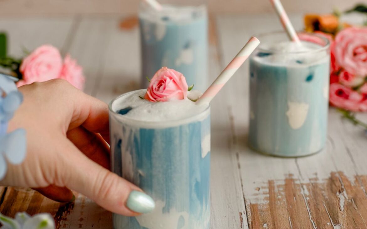 cover image butterfly pea flower latte close up womans hand holding blue latte with straw and pink rose bud as garnish with blue dress with butterfly sleeves