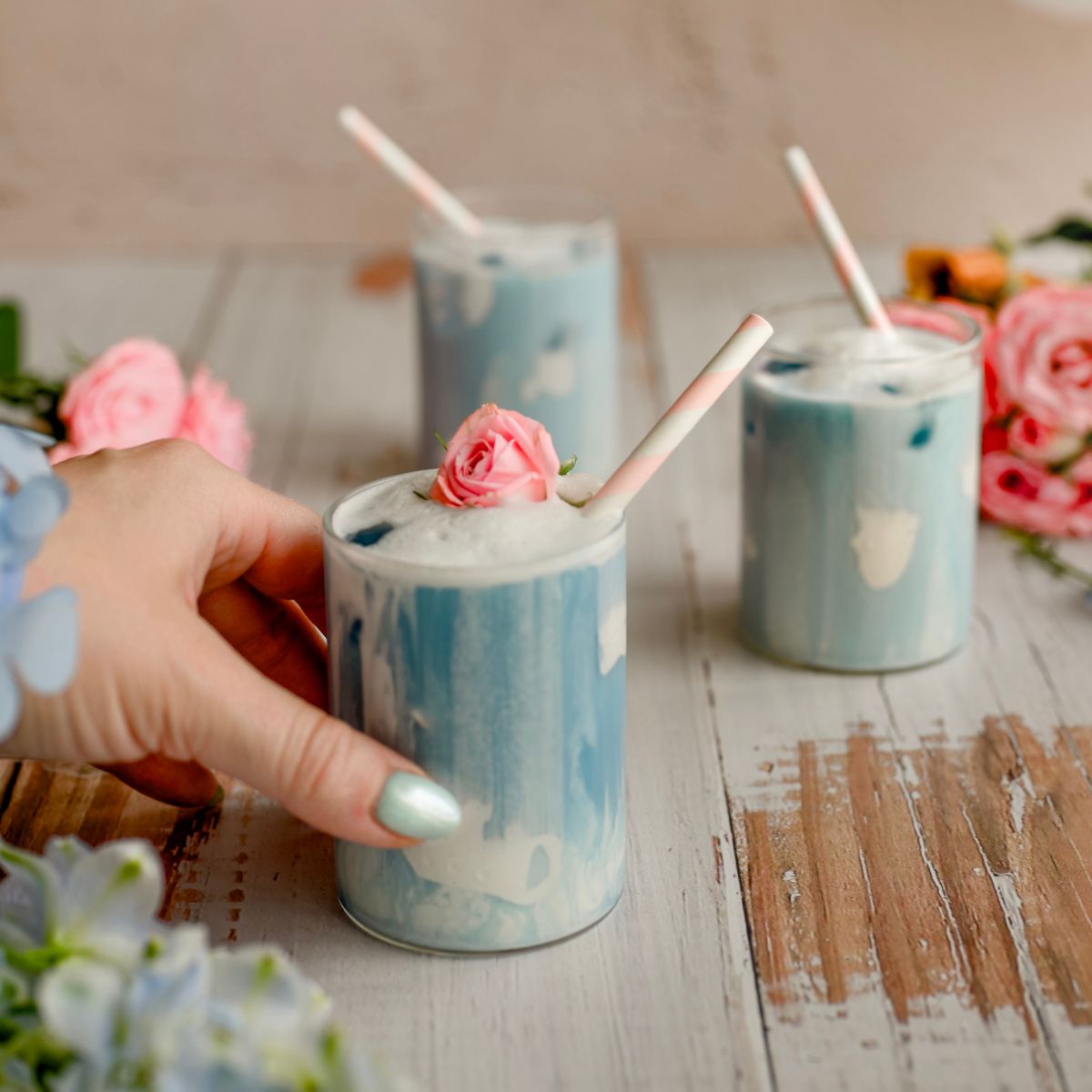 cover image butterfly pea flower latte close up womans hand holding blue latte with straw and pink rose bud as garnish with blue dress with butterfly sleeves