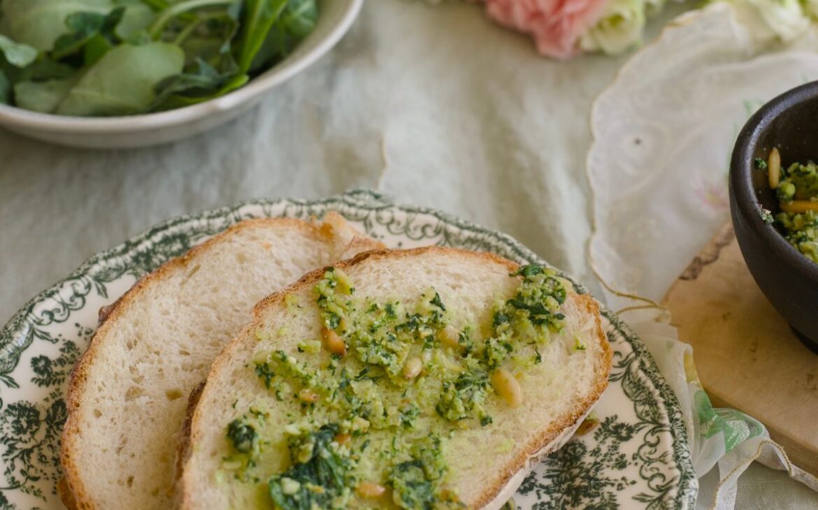 close up image of sliced sourdough bread with dandelion greens pesto next to a bowl of dandelion greens and mortar and pestle filled with pesto