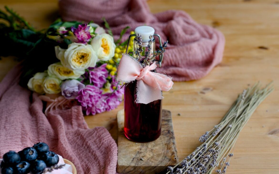 close up image of bottle with purple syrup containing blueberry lavender syrup on a rustic table in front of a bouquet of flowers and sprigs of lavender