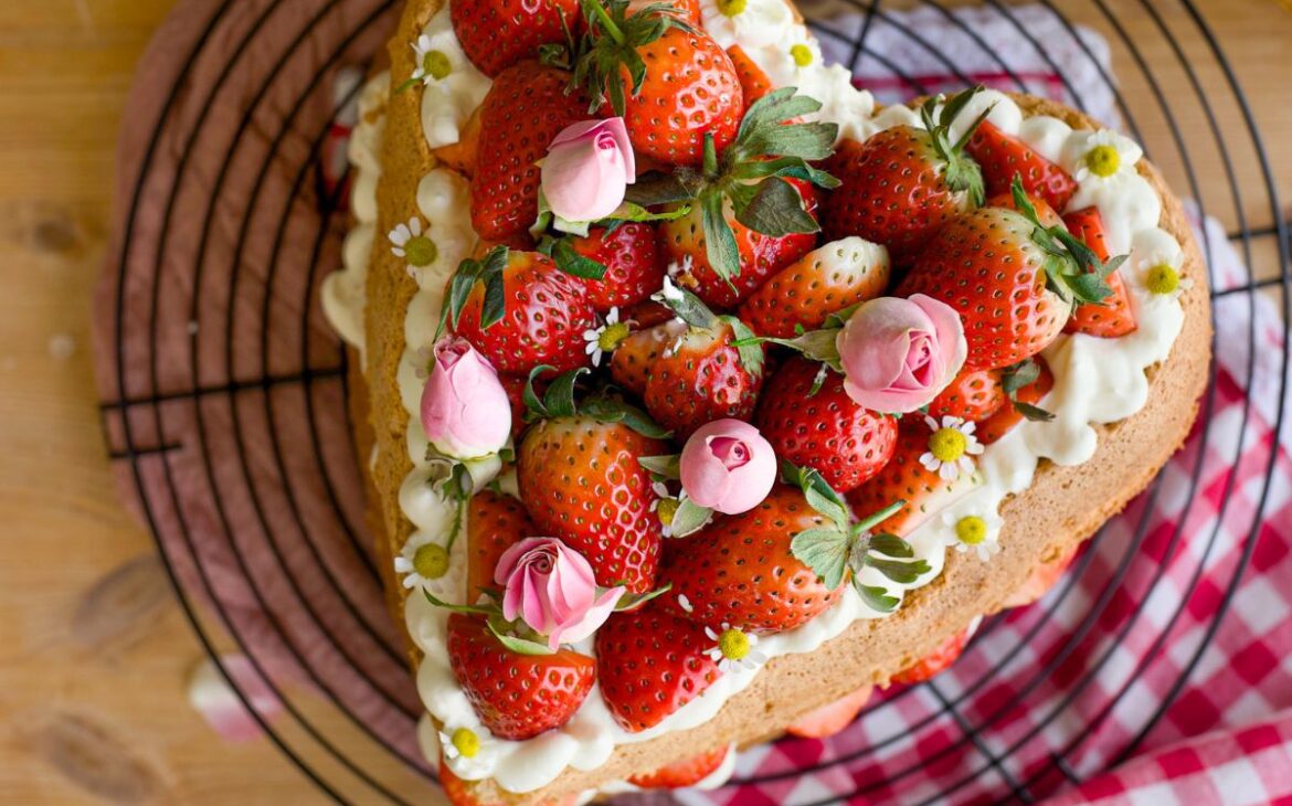 close up image of heart shaped strawberry sponge cake topped with chamomile whipped cream and strawberries and chamomile flowers and roses on top of a cooling rack with a layered gingham tea cloth on top of a wooden table