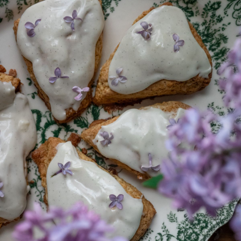 close up overhead image of scones with white glaze with vanilla bean specks garnished with dainty purple lilac flowers on a vintage green tray