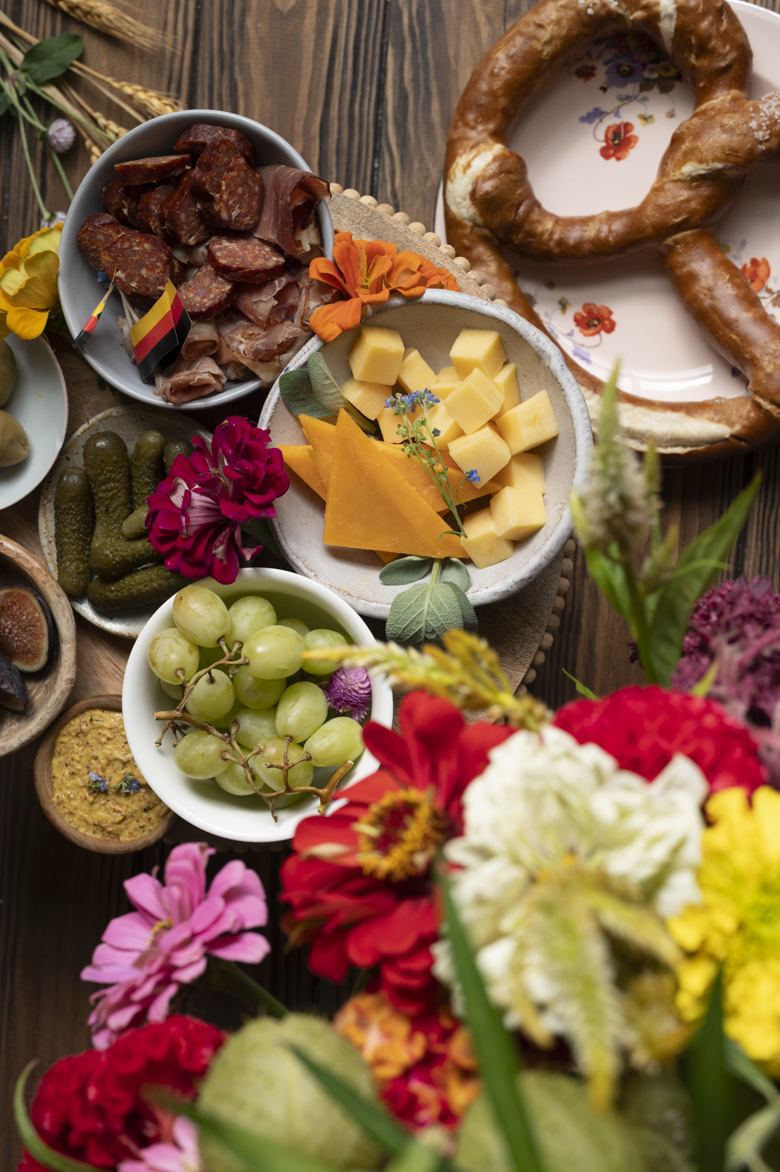 overhead shot on wooden background with small bowls of green grapes, sliced cheese, green olives, figs, and German meats and a soft pretzel for soft pretzel charcuterie board oktoberfest
