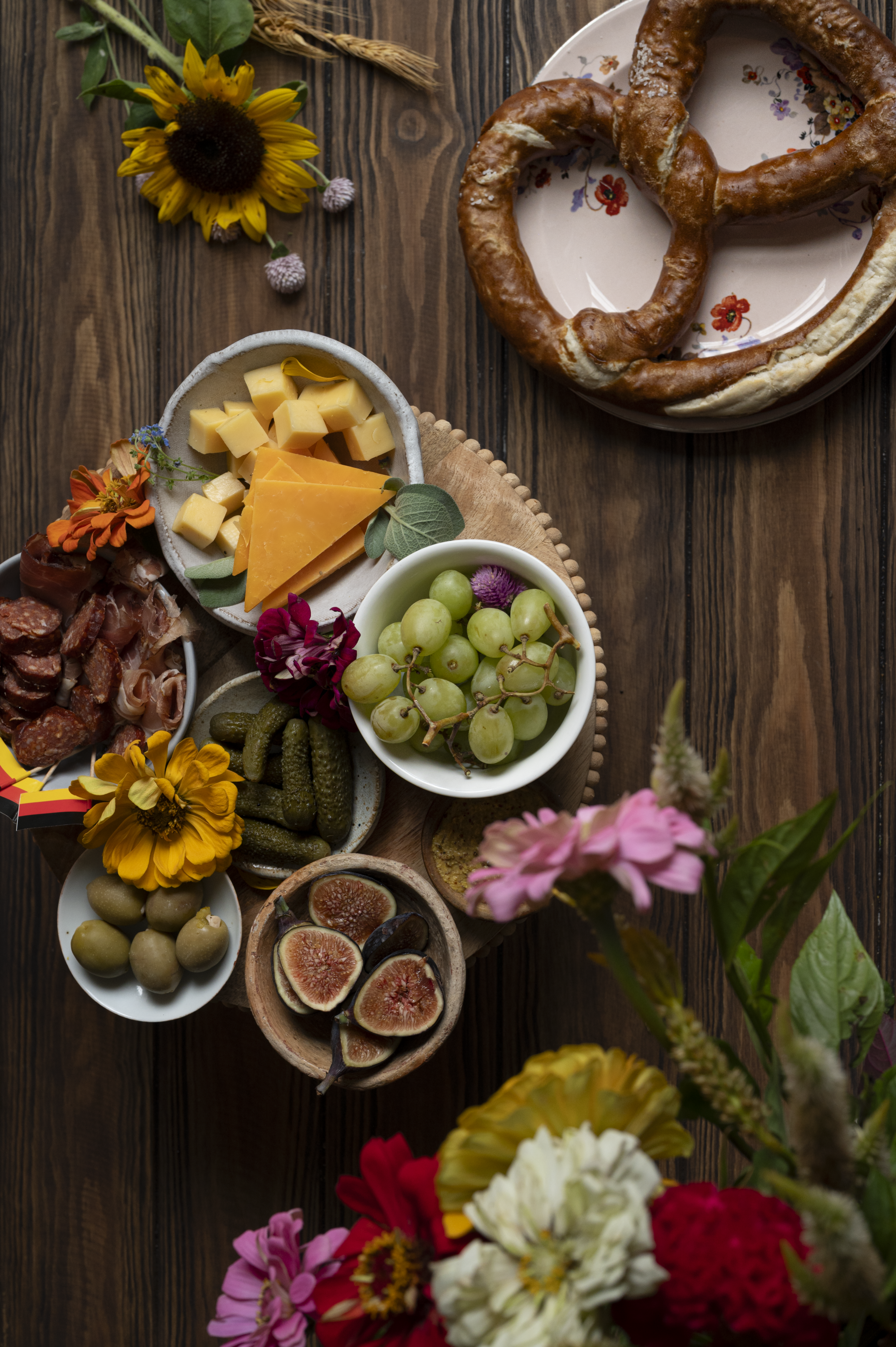 overhead shot on wooden background with small bowls of green grapes, sliced cheese, green olives, figs, and German meats and a soft pretzel for soft pretzel charcuterie board oktoberfest