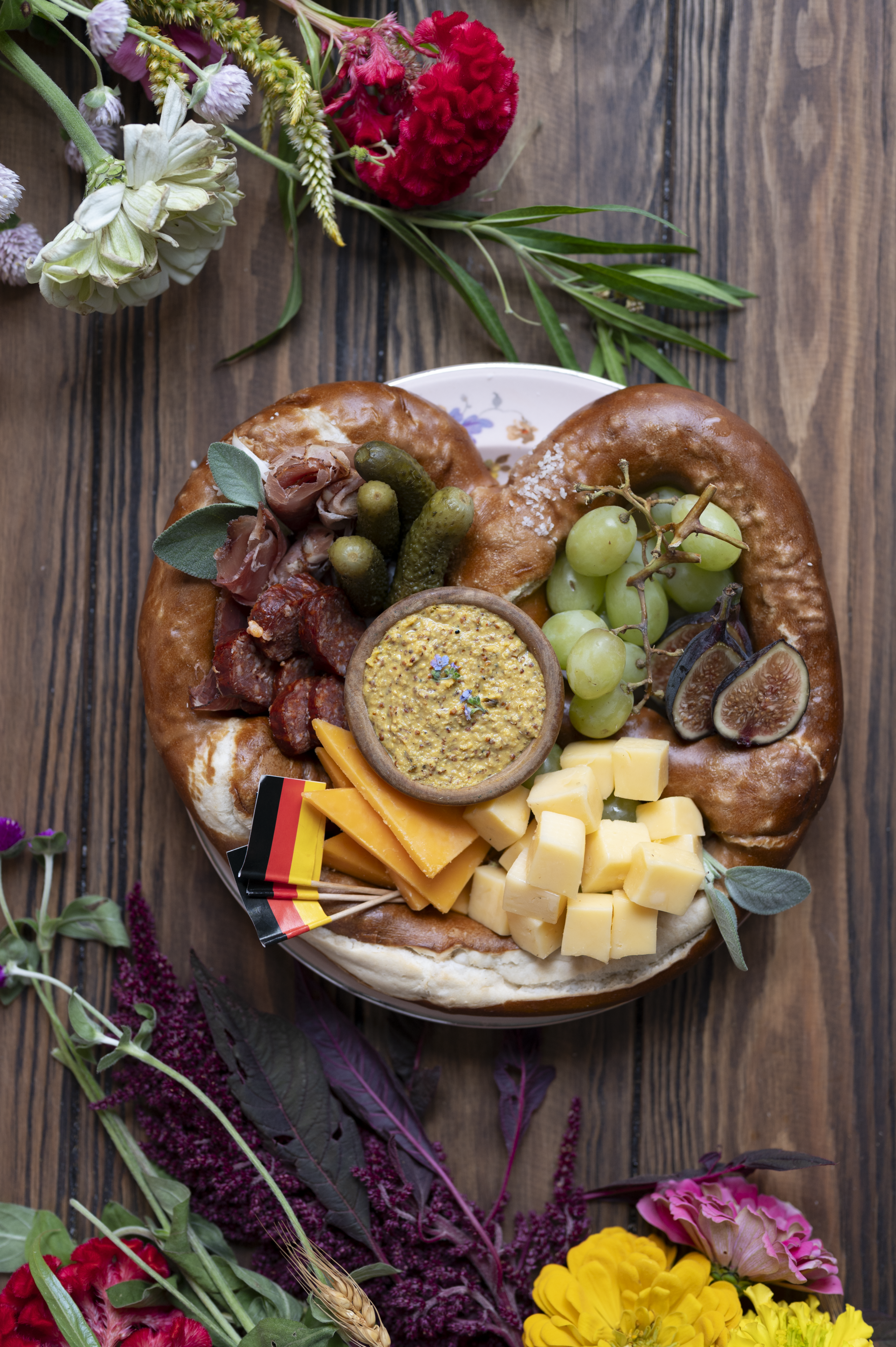 overhead shot of big Bavarian soft pretzel with fruit, meat and cheese for a soft pretzel charcuterie for Oktoberfest with mini Oktoberfest flag picks