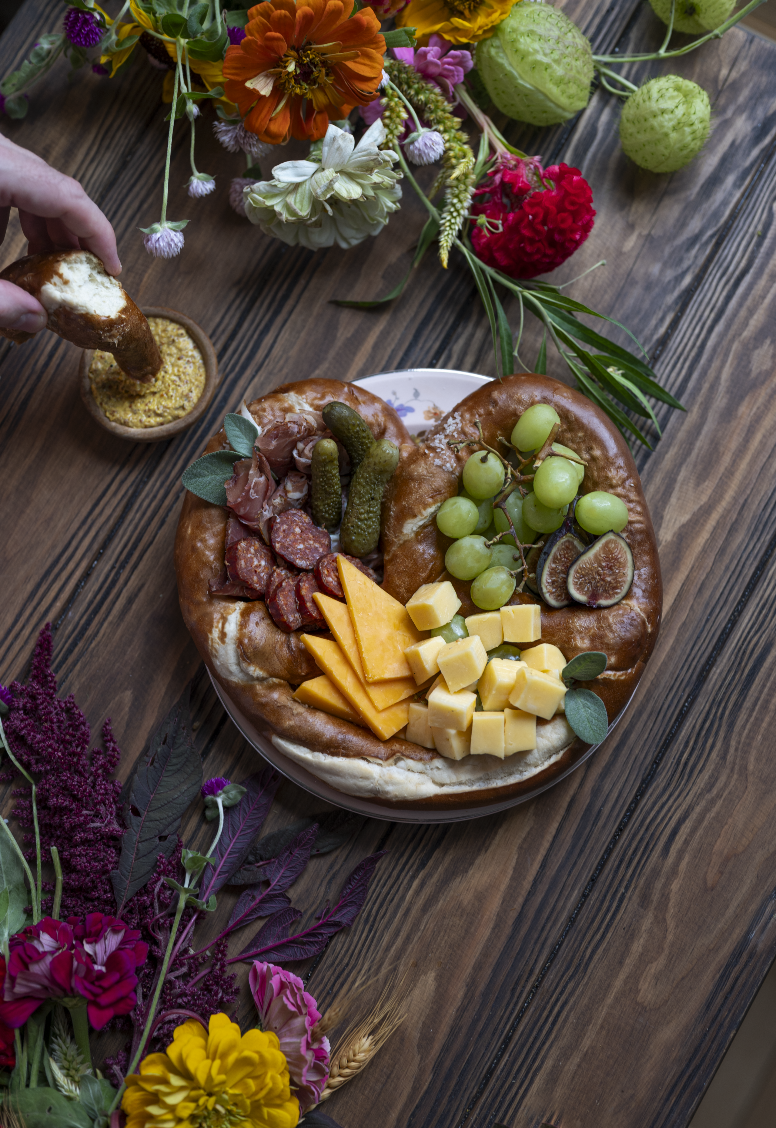 Overhead view of a salted pretzel surrounding whole-grain mustard, sliced salami, pickles, cheese, grapes and figs, with flowers around the platter.